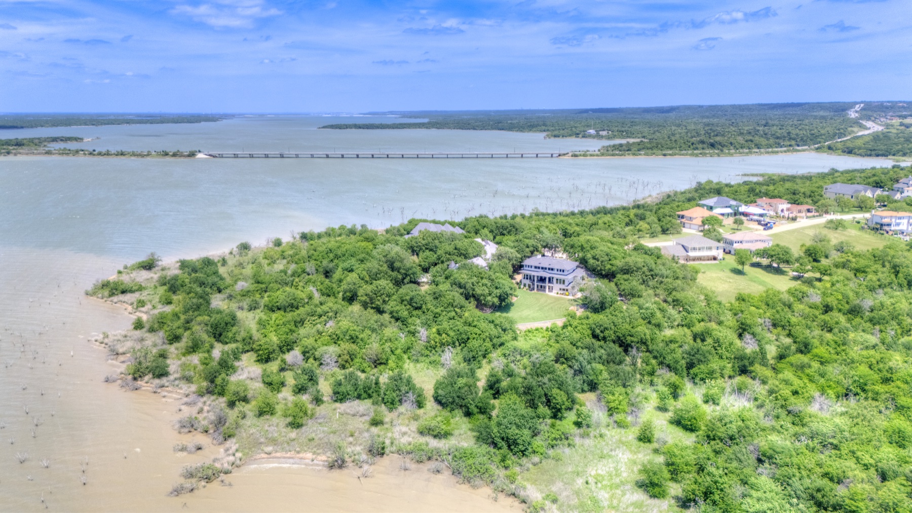Wide aerial drone view showing 1132 Mallard Pointe Drive sitting on a wooded peninsula surrounded by Joe Pool Lake stretching to the horizon, with the lake bridge crossing the upper portion of the frame.