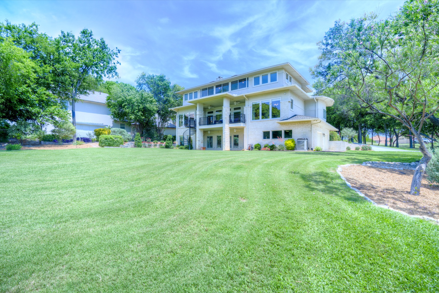 Rear elevation of the estate viewed from the back lawn: three levels with a multi-story covered balcony, large windows facing the lawn, the lawn rolling out toward the water beyond.