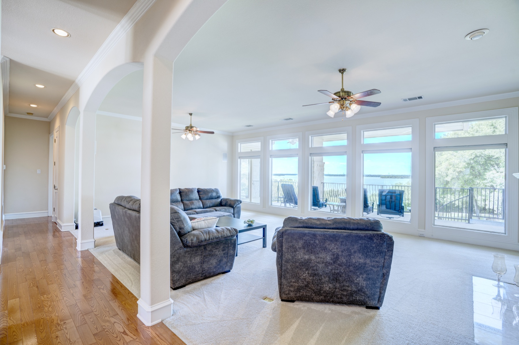 The great room interior, with floor-to-ceiling windows along the back wall framing a panoramic view of Joe Pool Lake. Pale stone flooring, neutral upholstery, high ceilings, mid-day light pouring across the room.