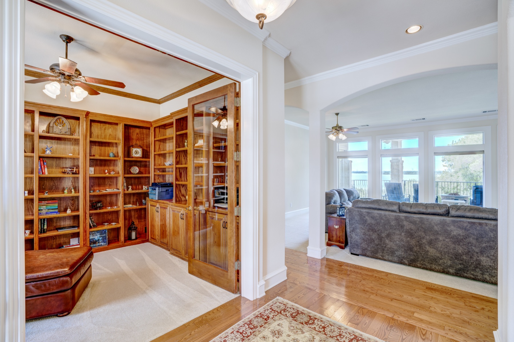 The library and study: floor-to-ceiling wood built-ins on two walls, a glass-paneled door to an adjoining room, hardwood flooring, an upholstered ottoman, view through to a sunlit great room beyond.