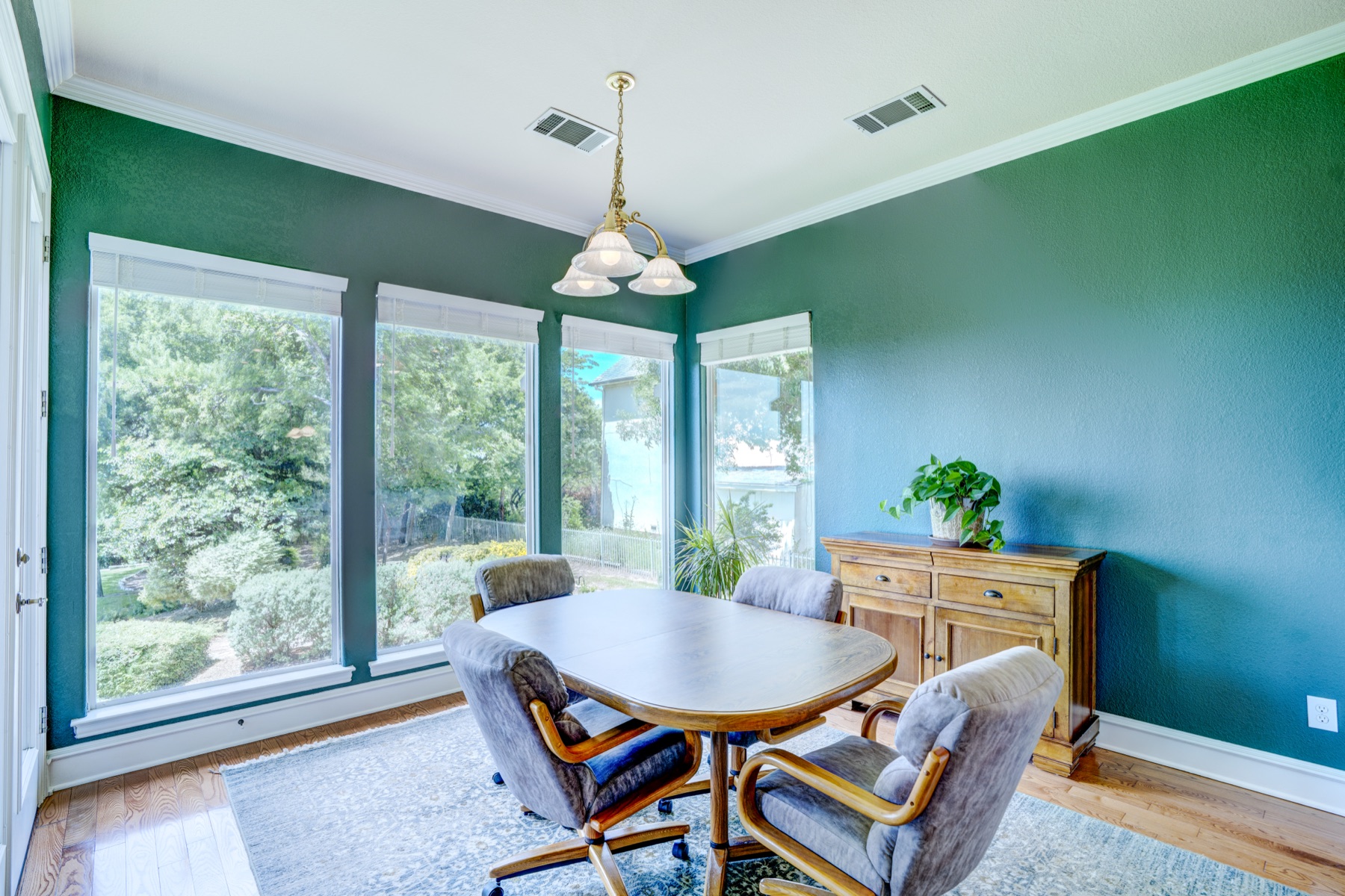 A dining room set in deep forest green, with a long oval table, four upholstered chairs, a brass chandelier overhead, and a wall of windows looking out to the property's mature landscaping.