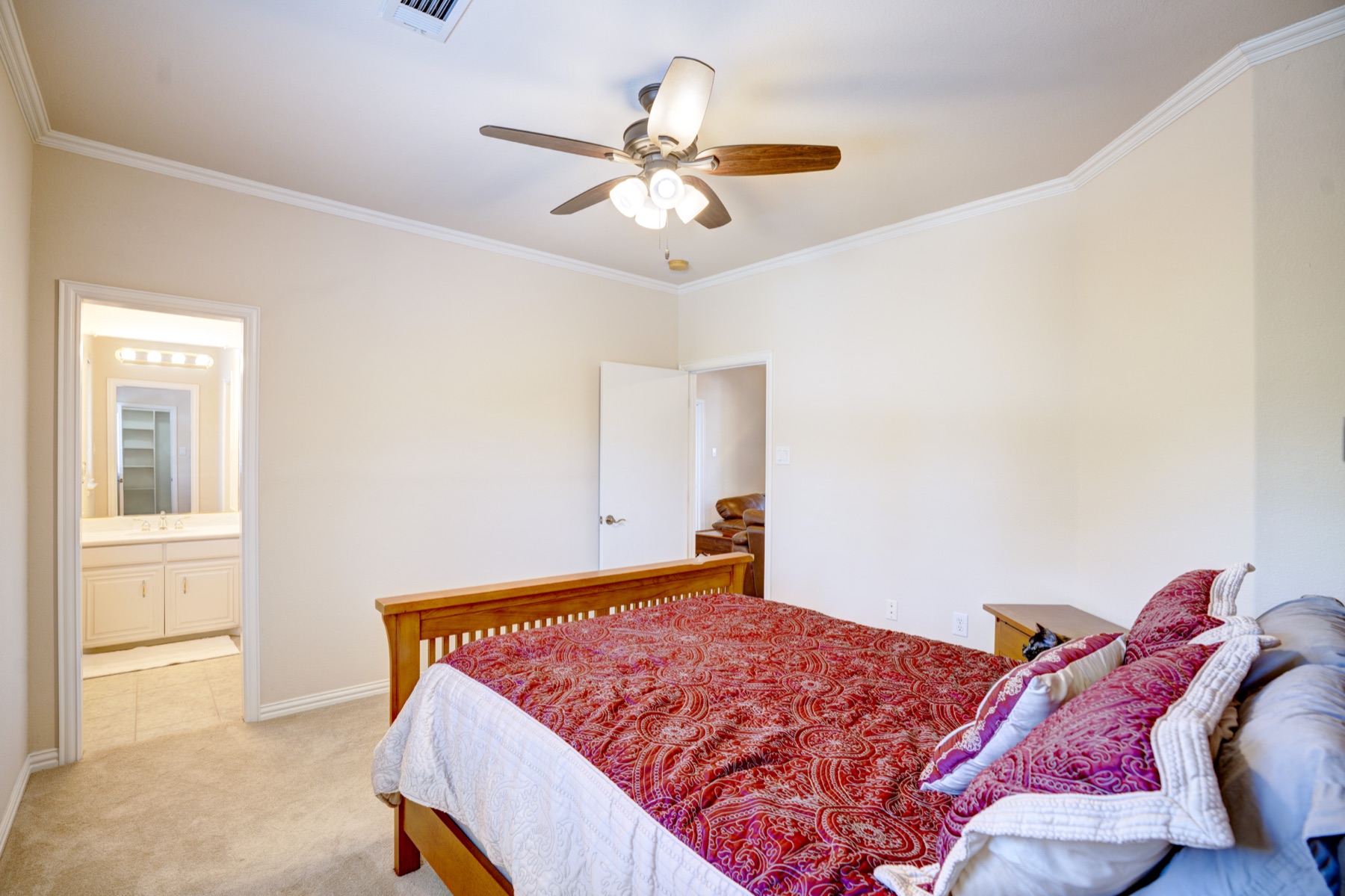 A bedroom suite with high ceilings, neutral walls, a wood sleigh-style bed framed by an open archway to an adjoining bath and walk-in closet, a ceiling fan overhead, soft natural light from a side window.