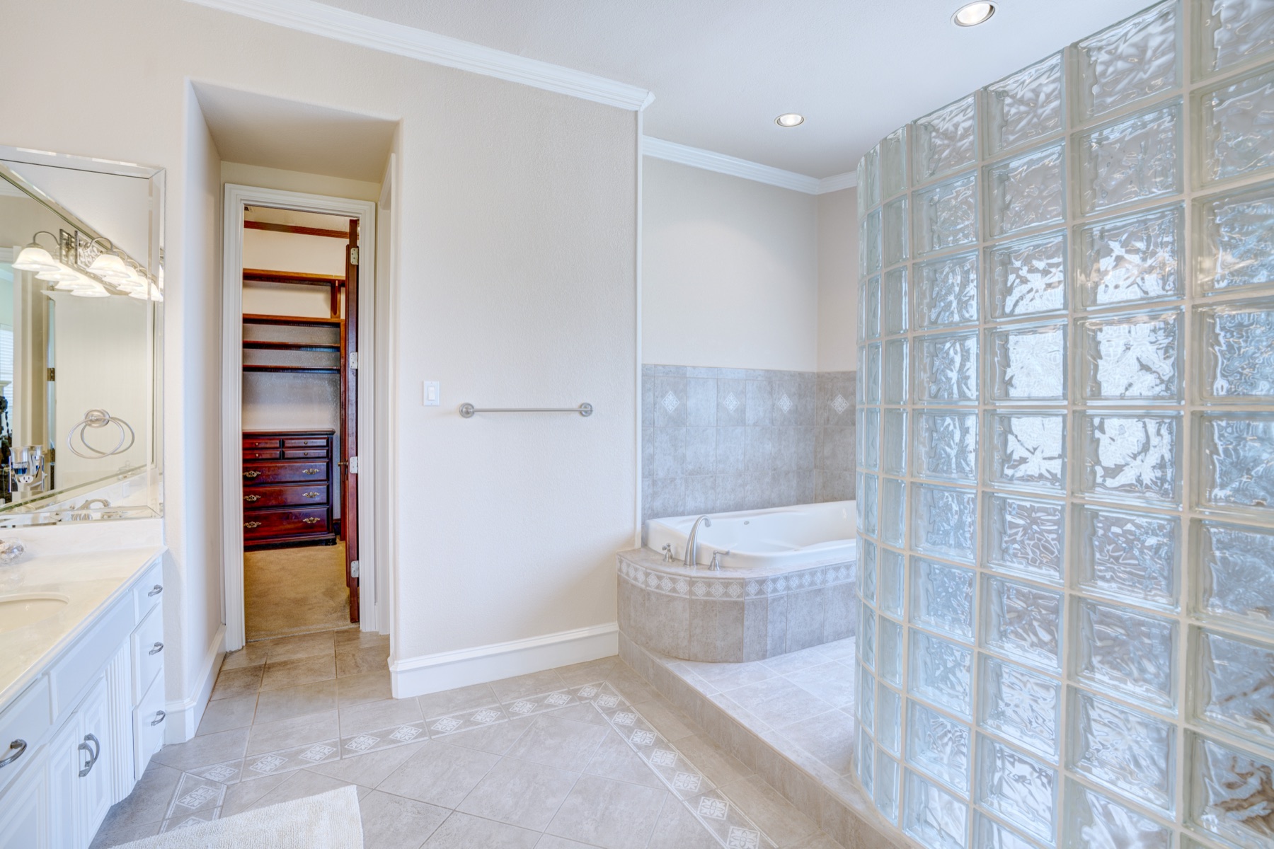 The primary bathroom: dual vanity along the left wall, a soaking tub set into a glass-block alcove, walk-in shower on the right, light marble flooring, oversized walk-in closet visible through a side opening.