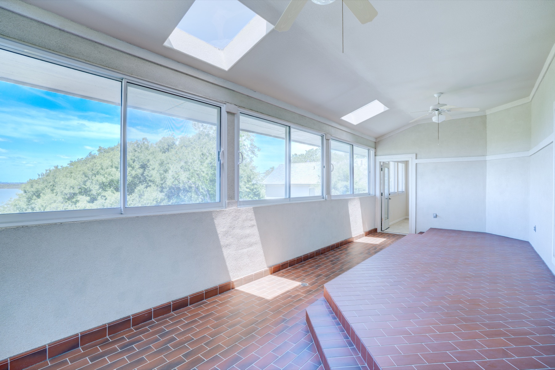 A second-floor sunroom or enclosed terrace with brick tile flooring, ribbon windows along three walls framing a panoramic view of Joe Pool Lake, skylights overhead, a quiet seating zone awaiting furnishing.