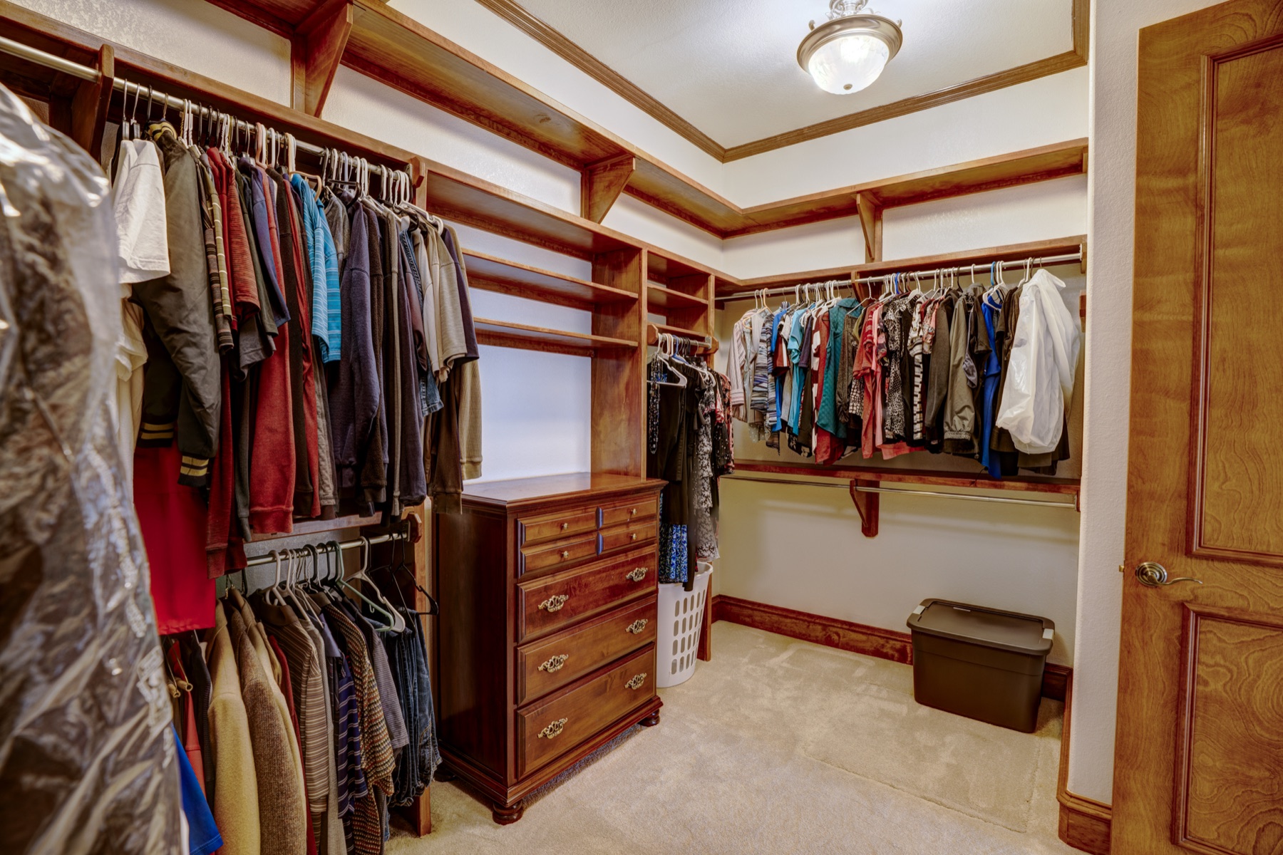 A walk-in closet with floor-to-ceiling wood built-ins, two parallel hanging rails of clothing, a built-in dresser at the center, recessed ceiling lighting, full-height ceiling and crown molding.