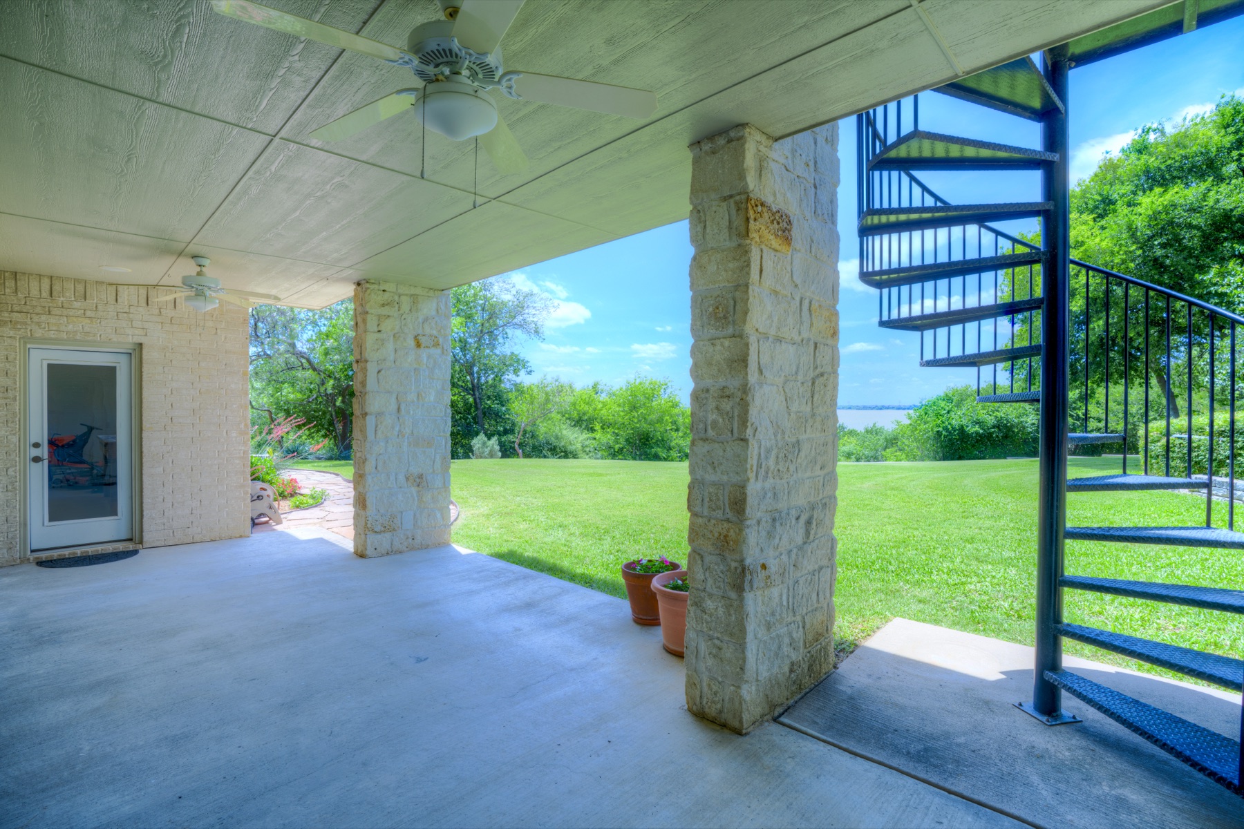 A covered outdoor patio area with stone columns, paver flooring, a wrought-iron spiral staircase ascending to the upper level on the right, the lawn and Joe Pool Lake visible in the distance through the columns.