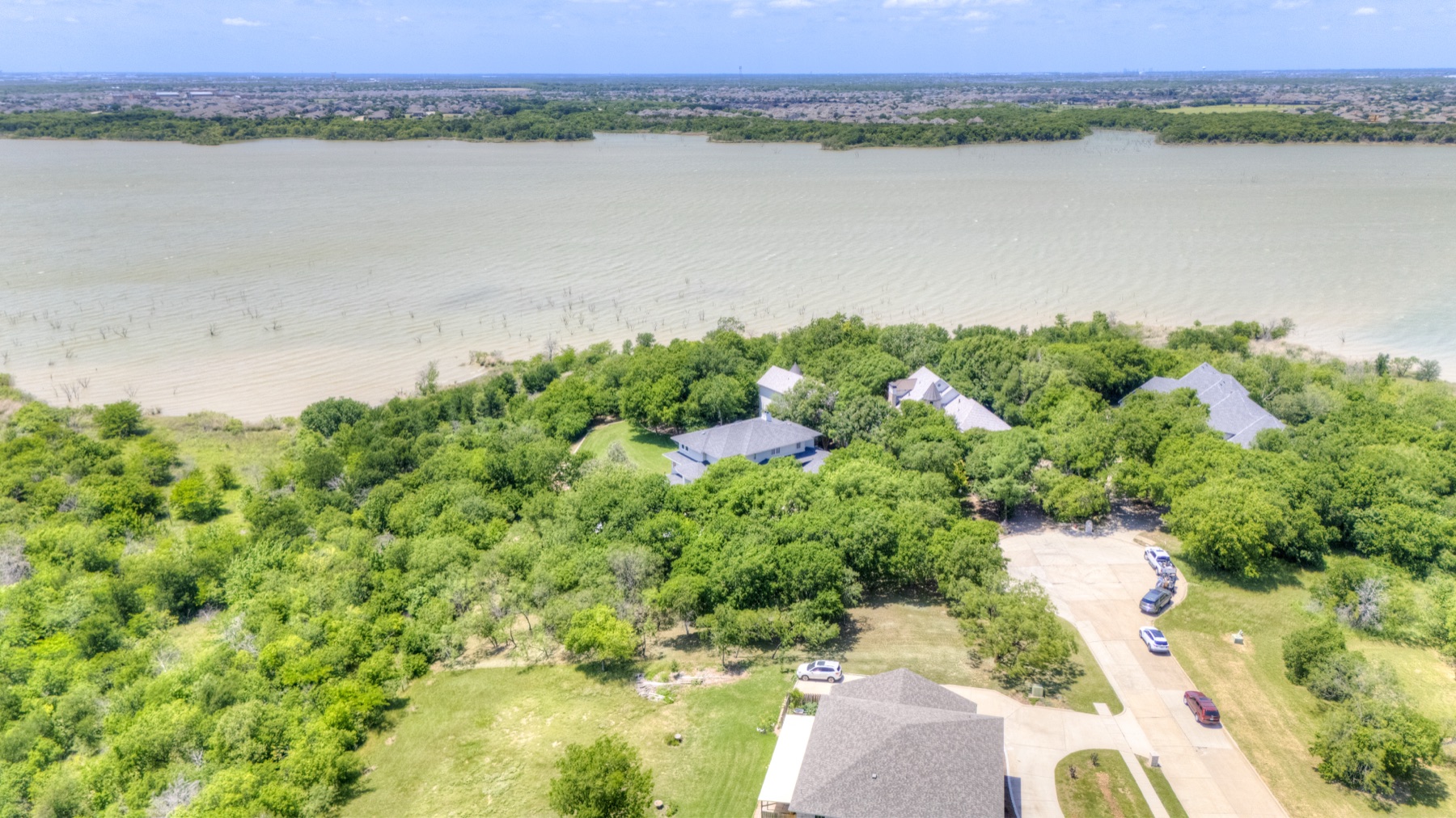 Cinematic wide aerial showing the full peninsula with the estate centered in mature trees, Joe Pool Lake stretching to the horizon on the left, the suburban grid of Cedar Hill visible in the far distance, the lake bridge crossing the top of the frame.